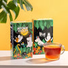 Two boxes of Moomin Jungle Joy tea with illustrated packaging, displayed on a white surface next to a glass mug filled with amber tea, against a yellow background with a green leaf in the corner.