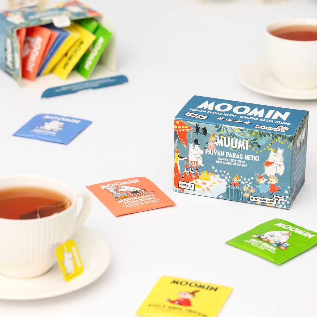 Moomin-themed tea bags and their colorful individual packets arranged on a white surface, with white teacups and saucers in the background and foreground. The box prominently shows the Moomin brand and character illustrations.