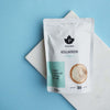A white pouch of Puhdistamo Collagen Natural by Puhdistamo rests on a white marble slab with a light blue background. The packaging displays a black leaf logo, Finnish text, and emphasizes its Bodybalance collagen peptide content.