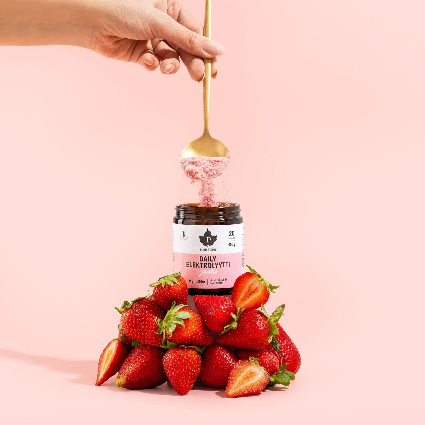 A hand holds a golden spoon above a jar of Puhdistamo Daily Electrolyte Powder Strawberry, surrounded by fresh strawberries against a pale pink background—showcasing daily hydration and natural strawberry flavor.
