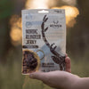 A hand holds a Renjer Nordic Reindeer Jerky Sea Salt pack outdoors, featuring reindeer antlers, "100% natural" text, and an image of sliced reindeer meat in a bowl.