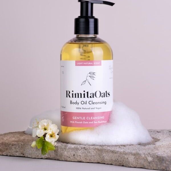 Rimita Green's RimitaOats Body Oil Cleansing, made with Finnish oats, rests on a stone slab with foam and white flowers. This 100% natural shower oil hydrates and gently cleanses your skin against a soft pink background.
