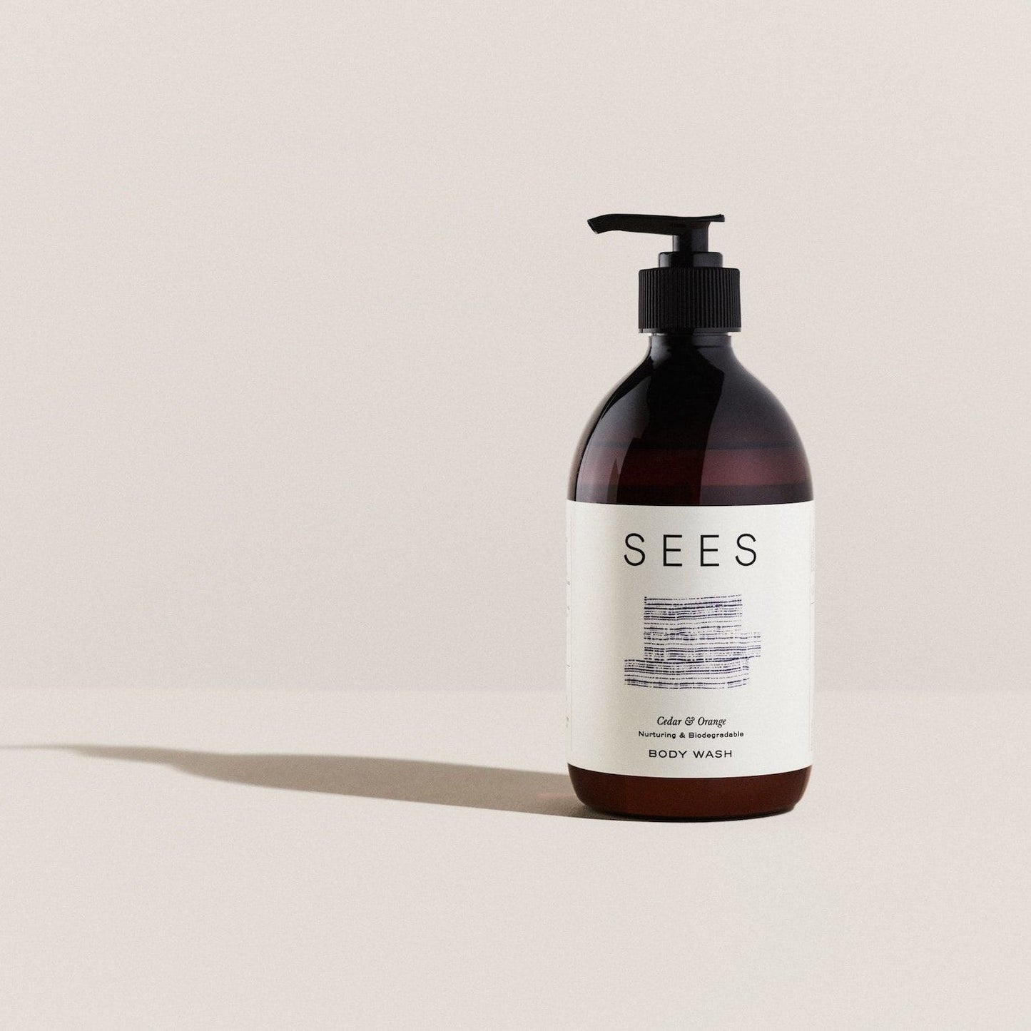 A brown pump bottle of SEES Company Body Wash, Cedar & Orange, stands on a light beige surface with a plain beige background. Its white label shows minimal black text and a blue abstract design, reflecting its plant-based cleanser formula.