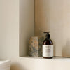 A brown pump bottle of SEES Company’s Cedar & Orange Body Wash rests on a minimalist bathroom shelf beside a stone block, with folded white towels nearby and light beige walls in the background.