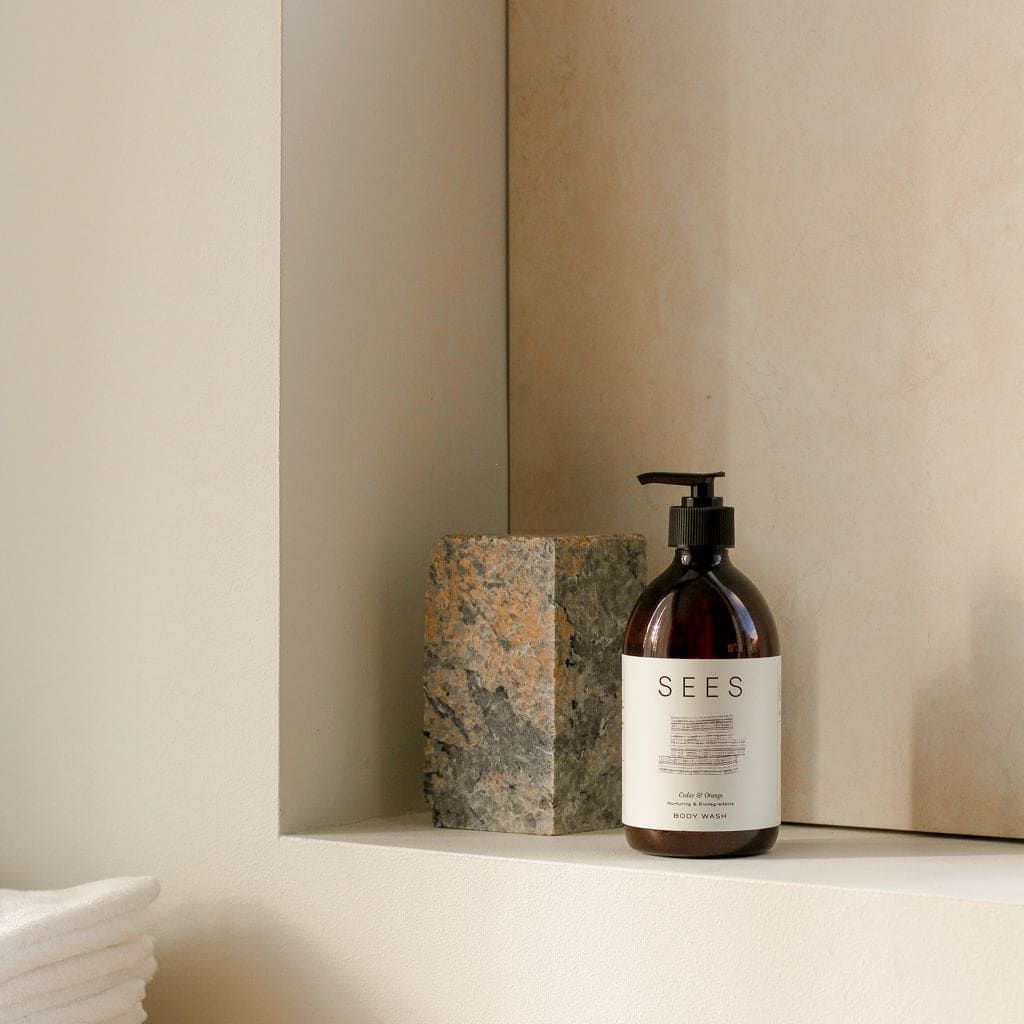 A brown pump bottle of SEES Company’s Cedar & Orange Body Wash rests on a minimalist bathroom shelf beside a stone block, with folded white towels nearby and light beige walls in the background.
