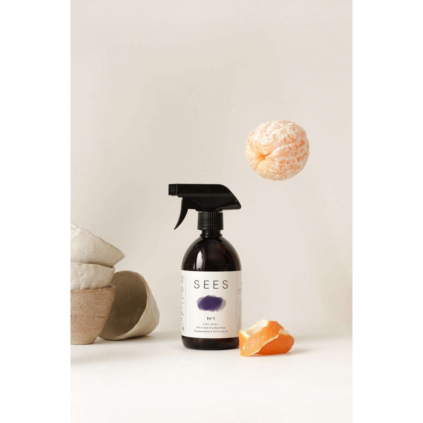 A dark SEES Company spray bottle labeled SEES Dishwashing Liquid stands upright by a peeled orange and ceramic bowls, with a whole mandarin floating above, highlighting our biodegradable, eco-friendly dish soap against a neutral background.