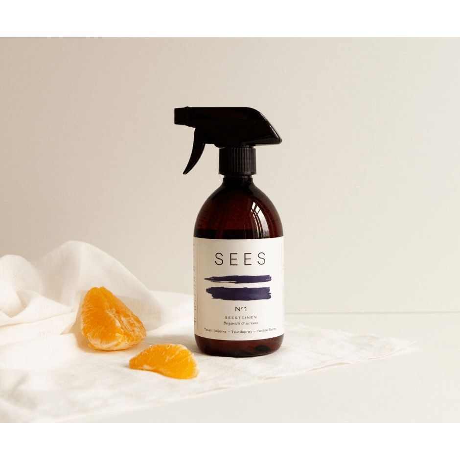 A brown SEES Company Textile Spray No. 1 Serene, Bergamot & Lemon bottle rests on a white cloth beside two orange tangerine segments, with a white background and soft lighting.