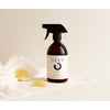 A dark brown spray bottle labeled SEES Textile Spray No. 2 Fresh, Orange & Peppermint by SEES Company stands on a white surface with a white cloth and two lemon wedges, set against a neutral, minimalistic background.