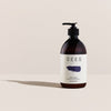 A dark amber pump bottle from SEES Company labeled "SEES Shampoo No. 1 Cedar & Orange" with a simple design, navy blue brushstroke, and the words Gel & Deep Shampoo on white stands upright on a neutral surface.
