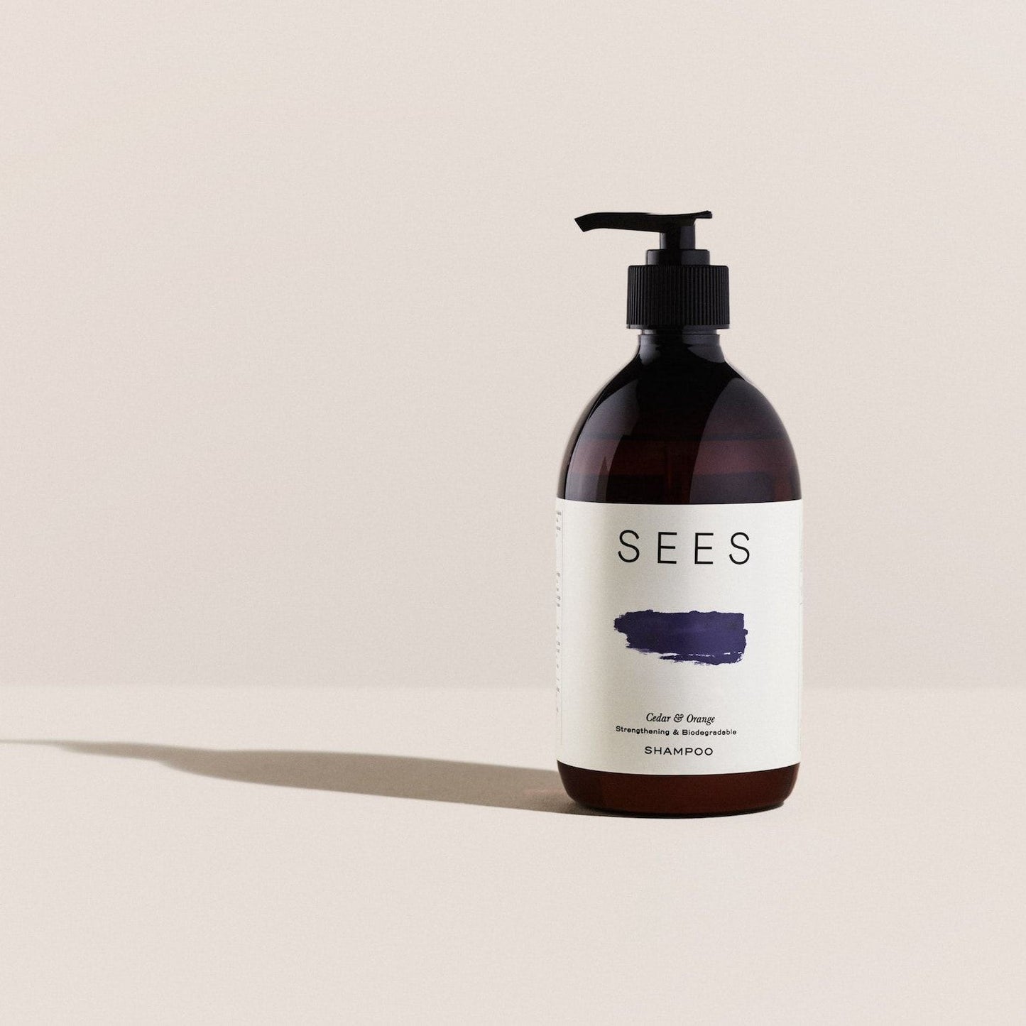 A dark amber pump bottle from SEES Company labeled "SEES Shampoo No. 1 Cedar & Orange" with a simple design, navy blue brushstroke, and the words Gel & Deep Shampoo on white stands upright on a neutral surface.