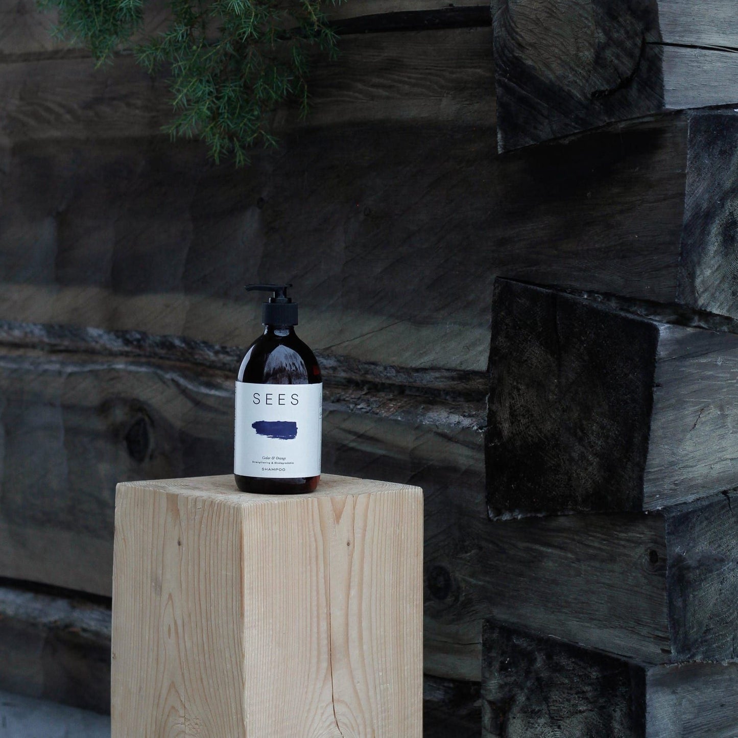 A bottle of SEES Company Shampoo No. 1 Cedar & Orange, made with essential oils, sits on a light wooden block before a rustic log wall and hanging greenery.