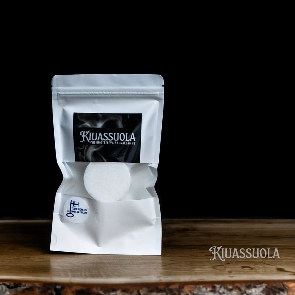 Ein weißer, wiederverschließbarer Beutel mit Kiuassuola-Sauna-Salz steht auf einer Holzfläche vor einem schwarzen Hintergrund. Davor befindet sich eine runde weiße Saunasalztablette, auf der das Kiuassuola-Logo in der rechten unteren Ecke zu sehen ist.