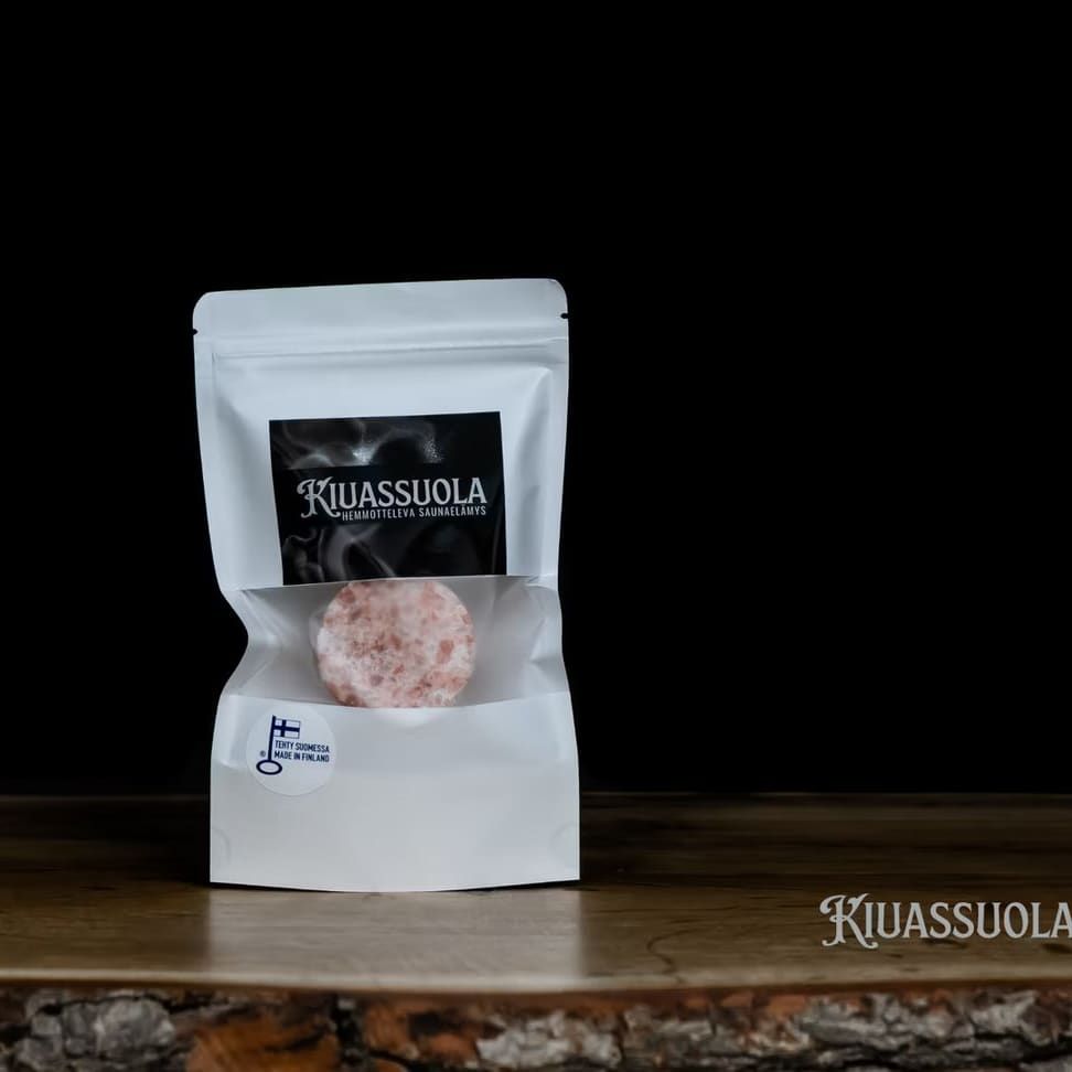 Ein weißer wiederverschließbarer Beutel mit der Aufschrift "Sauna Salt" der Marke Kiuassuola enthält ein rundes Himalaya-Kristallsalz. Die Verpackung ruht auf Holz vor einem schwarzen Hintergrund, mit "Kiuassuola" in der unteren rechten Ecke.