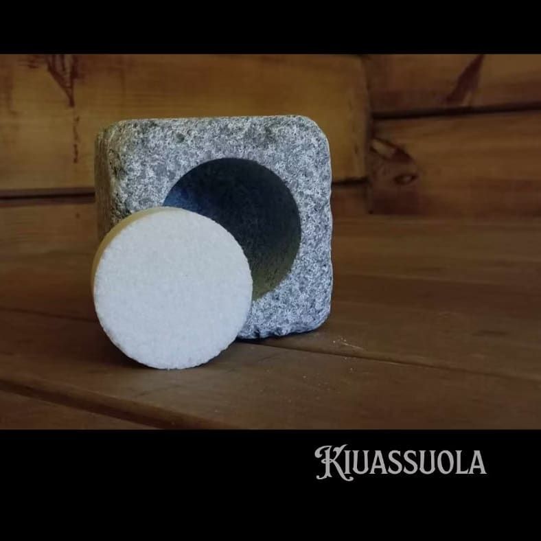 A round white sauna salt tablet from the Kiuassuola Sauna Salt ILMARI Starter Pack sits before a square stone holder with a circular hollow on wood, capturing the essence of wellness. The brand name "Kiuassuola" appears at the bottom right.