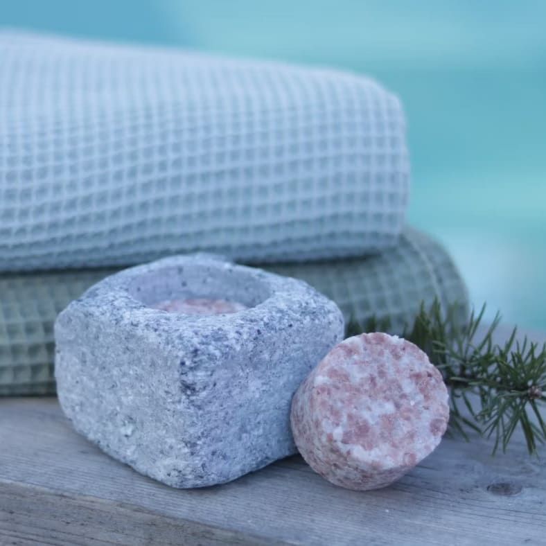 The Kiuassuola Sauna Salt ILMARI Starter Pack features a gray stone holder with natural sauna salt on wood. Light blue and green waffle towels and a pine branch in the background create a serene wellness vibe.