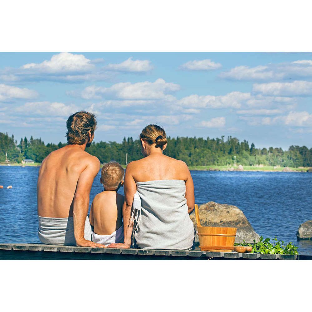 A man, woman, and child sit on a dock by a lake under a blue sky, wrapped in towels beside an Emendo Romantic Rose Sauna Honey jar, enjoying a serene retreat surrounded by trees and the moisturizing scent of roses.