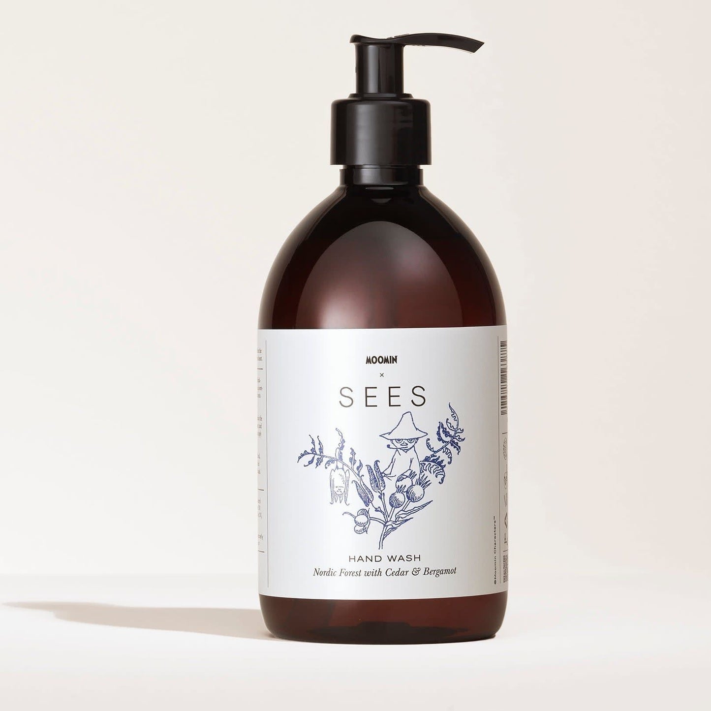 A brown pump bottle of SEES Company’s SEES x Moomin Hand Wash, featuring botanical illustrations and labeled “Nettle Forest with Cedar & Bergamot,” is displayed on a white background.