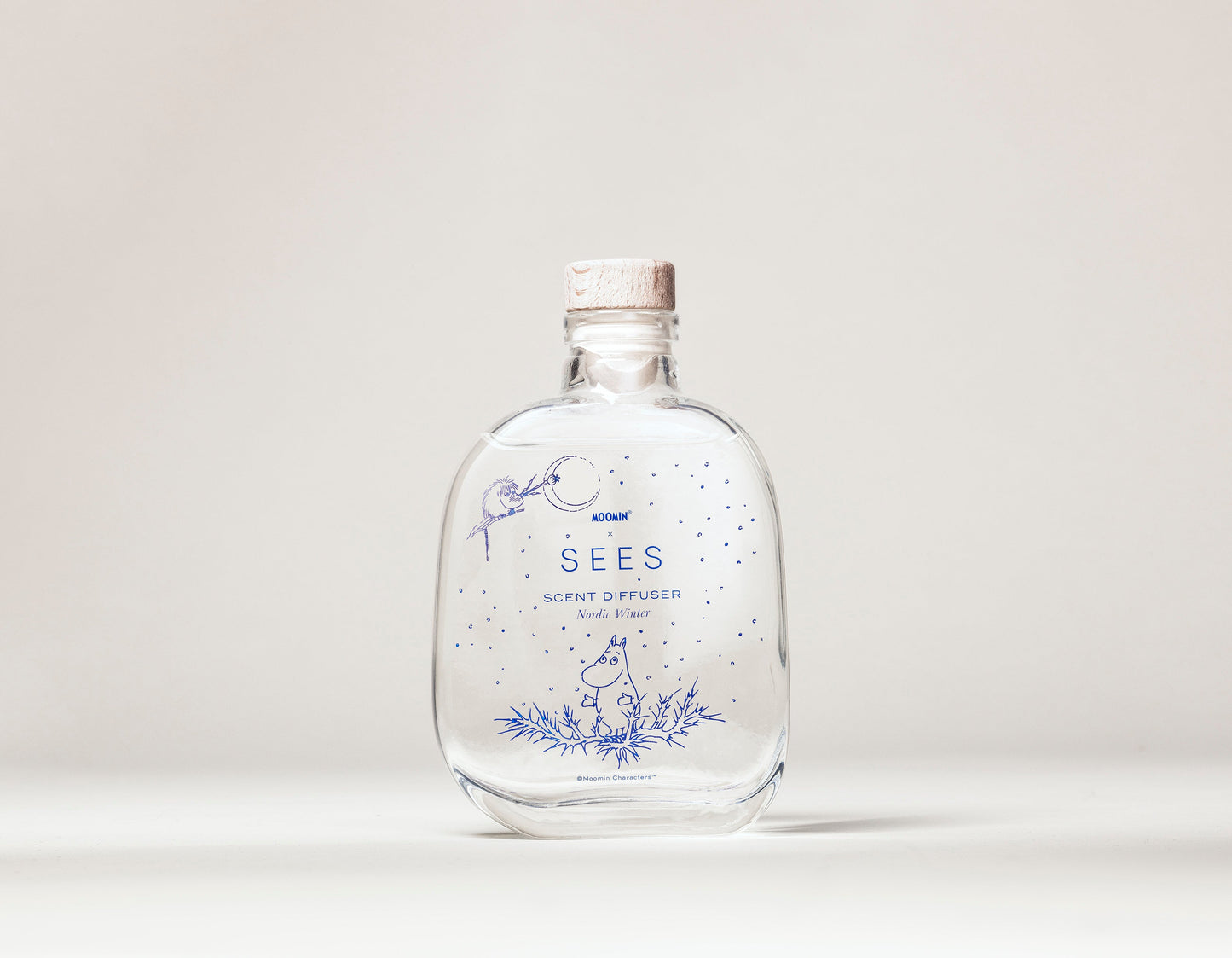 A clear glass bottle with a cork stopper labeled SEES x Moomin Room Diffuser Nordic Winter by SEES Company, featuring minimalist botanical and animal illustrations inspired by Finnish design, set on a plain light background.