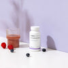 A white bottle of THERA Nordic Optimized Enzymes stands on a white surface next to a glass of pink liquid, with raspberries and blueberries nearby, suggesting digestive enzyme relief against a light purple background.