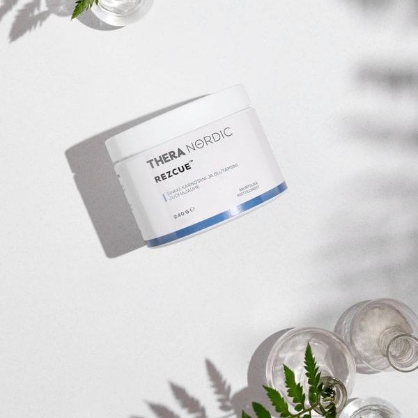 A white container labeled THERA Nordic Rezcue Drink Powder by THERA Nordic, designed for gut healing, sits on a white surface surrounded by glass vases with green fern leaves. Sunlight streams in, casting soft shadows.