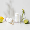 Two THERA Nordic Triple HCL bottles from THERA Nordic—one upright and one tipped with tablets spilling out—are shown with a lemon, halved lime, and small yellow flower, highlighting this digestive health supplement on a light background.