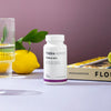 A bottle of THERA Nordic Triple HCL by THERA Nordic sits beside a glass of water, a lemon, a lime wedge, green leaves, and a yellow-covered book on a purple background.
