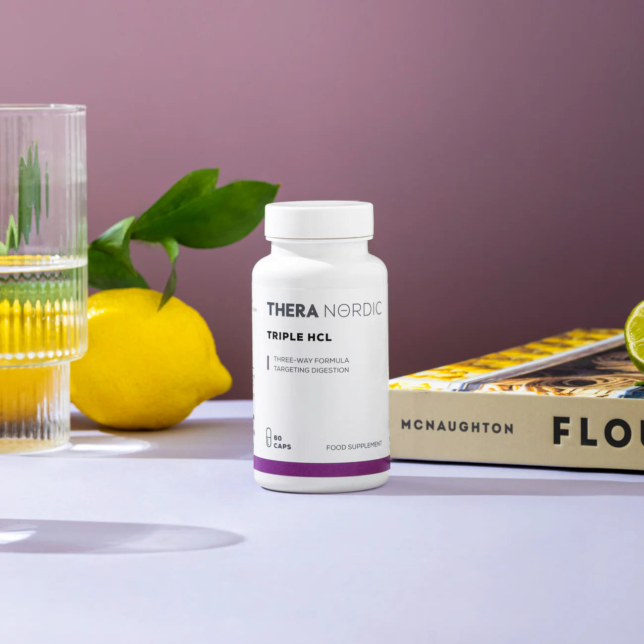 A bottle of THERA Nordic Triple HCL by THERA Nordic sits beside a glass of water, a lemon, a lime wedge, green leaves, and a yellow-covered book on a purple background.