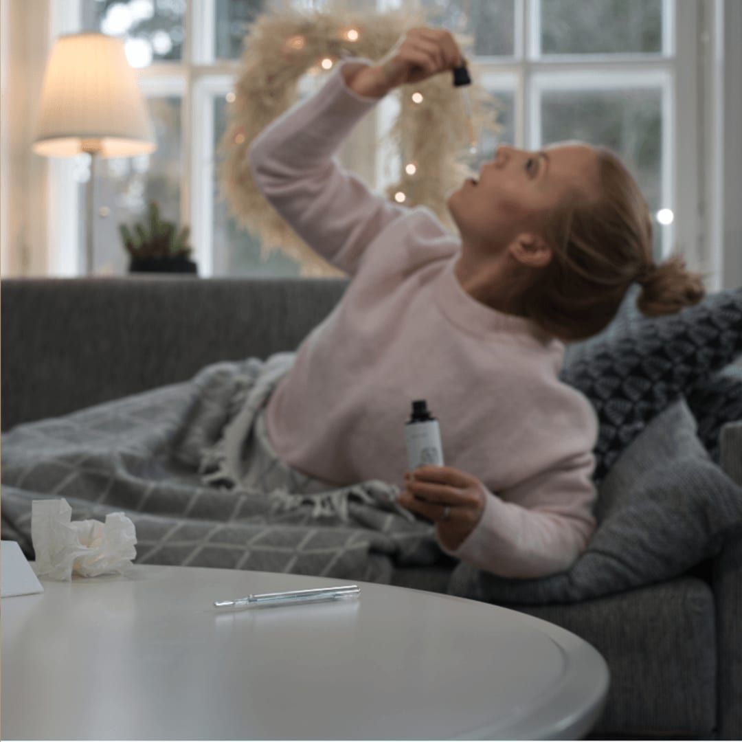 A woman lies on a couch under a blanket applying eye drops, with tissues and a thermometer nearby—she may be unwell and could benefit from immune support such as KÄÄPÄ Mushrooms Organic Shiitake Extract.