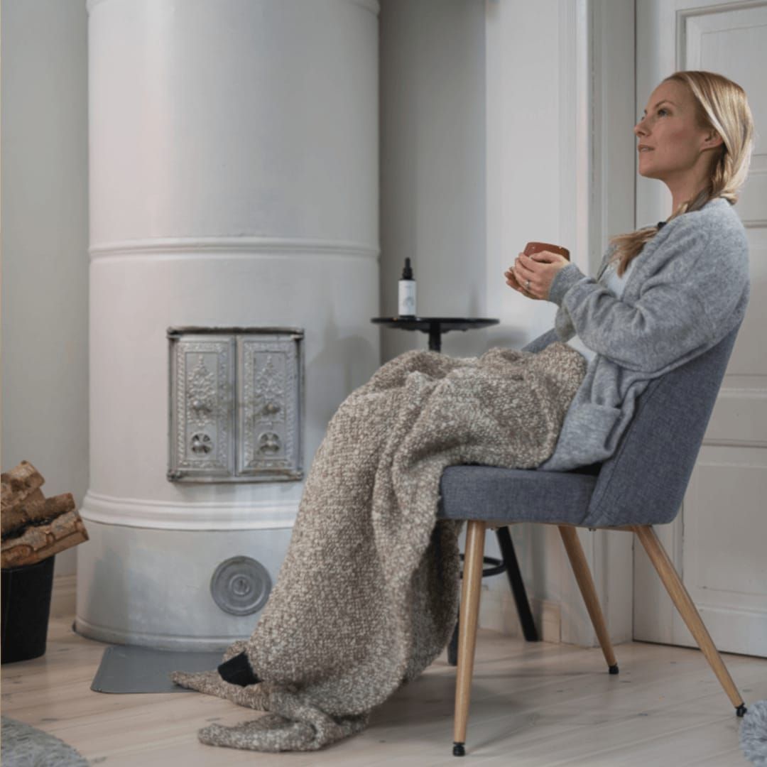 A woman sits in a gray chair, wrapped in a cozy blanket and holding a cup featuring KÄÄPÄ Mushrooms Reishi Extract. She faces a white fireplace with firewood stacked nearby in a modern, minimalist room.