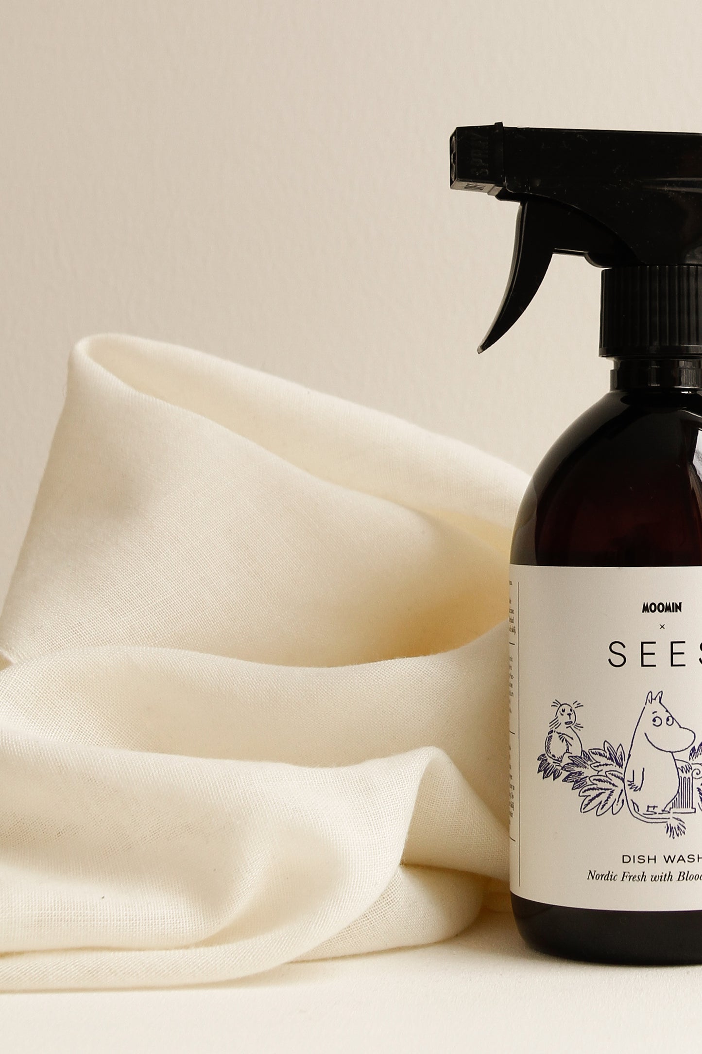 Ein SEES x Moomin Geschirrspülspray von SEES Company mit einer braunen Flasche und einem Etikett mit Moomin-Motiv steht neben einem weichen weißen Tuch vor einem hellen, neutralen Hintergrund.