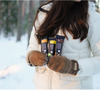 Eine Frau in einem weißen Wintermantel und braunen Fäustlingen steht in einem verschneiten Wald und hält drei Foodin Collagen Protein Bars in der Hand, darunter die Geschmacksrichtung Crunchy Caramel Cinnamon.