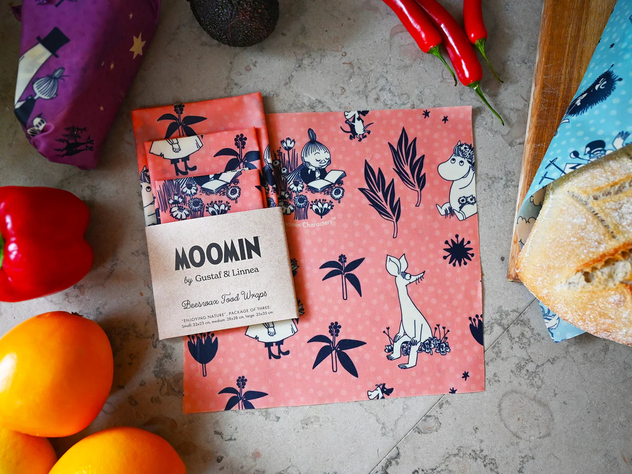 Die Gustaf & Linnea Moomin Bienenwachs-Lebensmittelpackung, eine umweltfreundliche, quadratische rosa Packung mit Mumin-Figuren, liegt auf einer Arbeitsplatte zwischen Orangen, Brot, Paprika und anderen Lebensmittelpackungen und wird von einem braunen Papieretikett gekrönt.