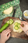 Zwei Hände legen Gurkenscheiben auf ein mit Käse belegtes Brot auf einem Holzbrett. Eine Gurke, ein Käselaib, ein Sparschäler und eine Schüssel mit Käsescheiben stehen daneben. Im Hintergrund sorgt eine Gustaf & Linnea Mumin Bienenwachs-Lebensmittelpackung für einen grünen Touch.