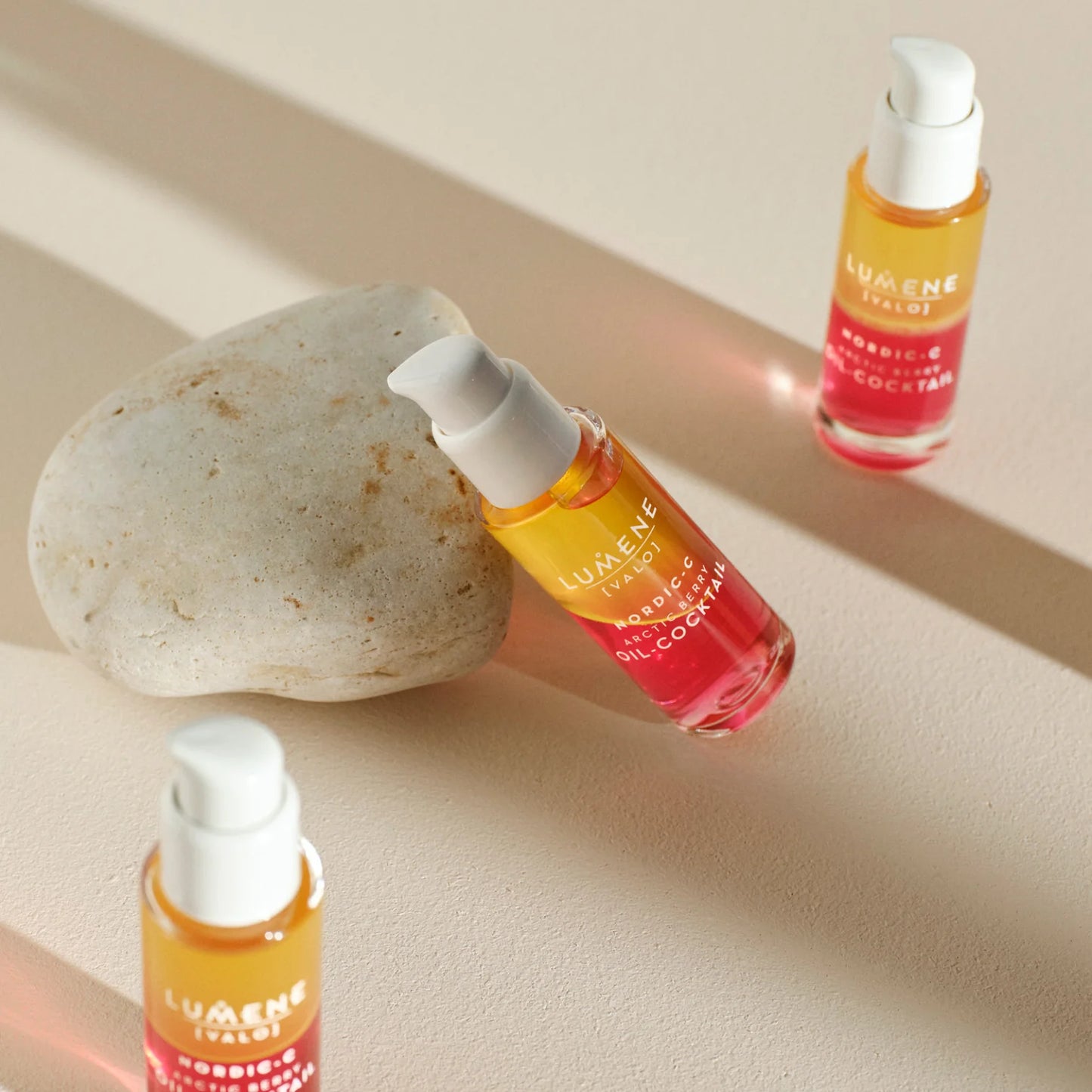 Drei Flaschen Lumene Nordic-C Arctic Berry Oil-Cocktail stehen auf einer beigen Fläche, eine lehnt an einem glatten Stein. Das orange-rosa schillernde Glas und die weißen Verschlüsse erinnern an das Leuchten der Haut durch den Extrakt der arktischen Moltebeere.