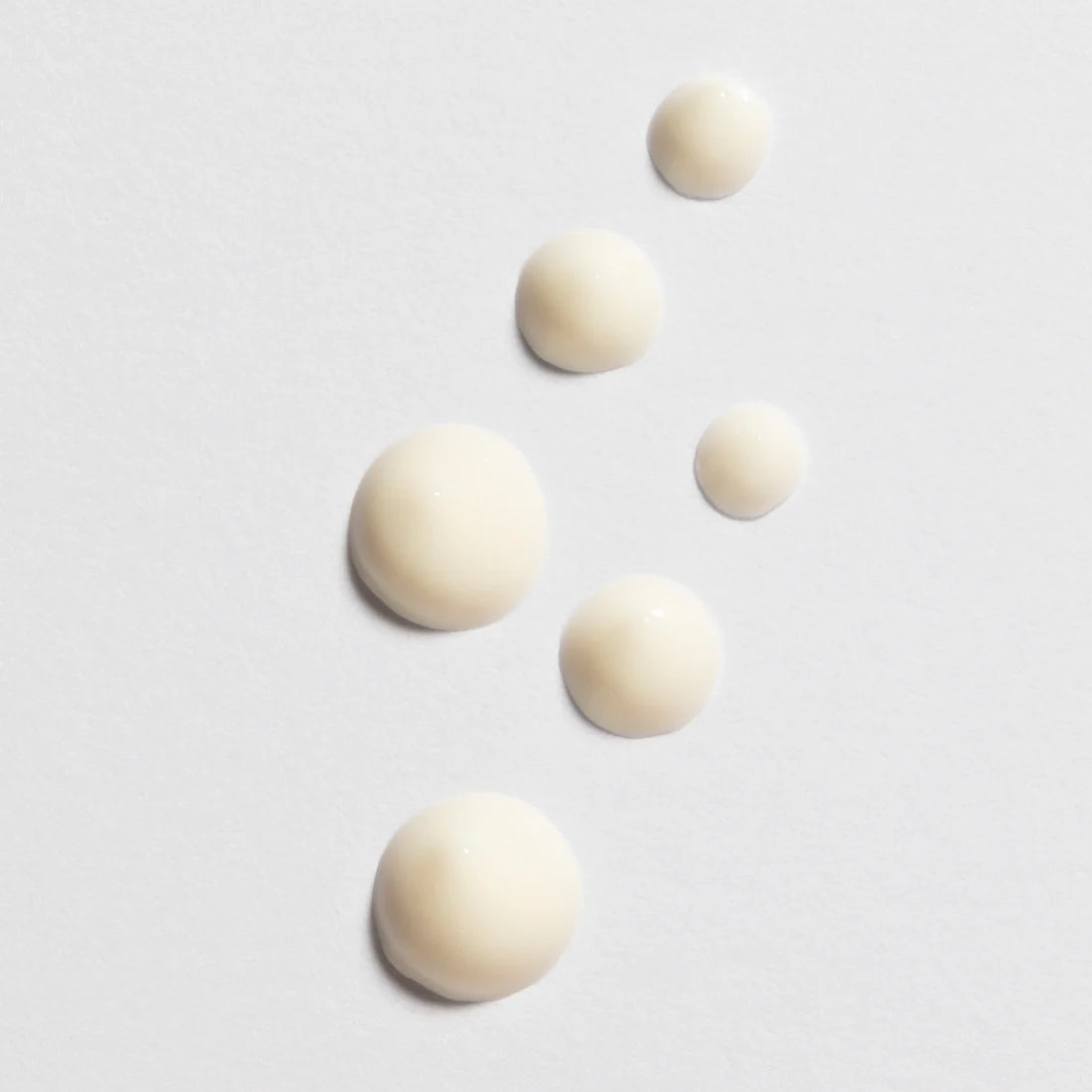 Sechs Tropfen der Lumene Nordic-C Glow Awakening Eye Cream von Lumene, einer cremigen, cremefarbenen Vitamin-C-Formel, sind diagonal auf einer weißen Fläche angeordnet, wobei die Tropfengröße von groß nach klein abnimmt.