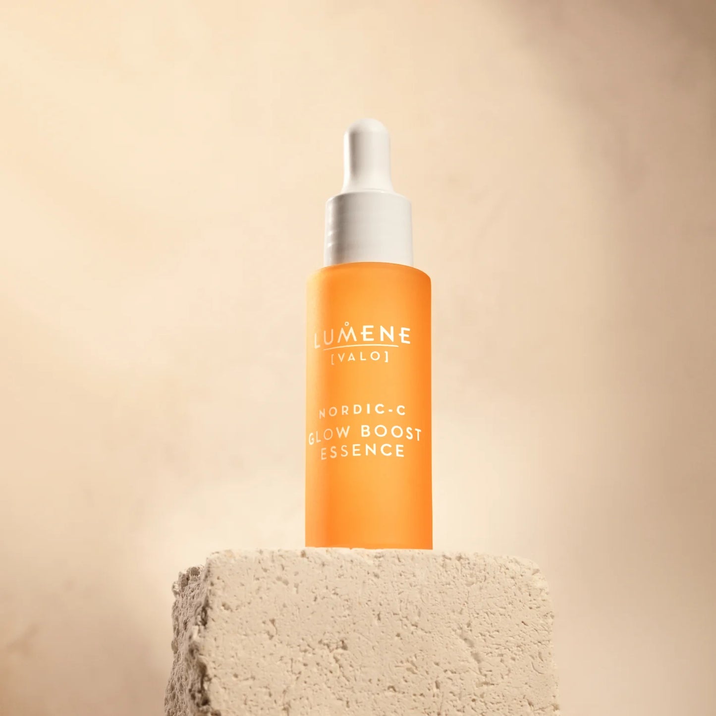 Ein Fläschchen Lumene Nordic-C Glow Boost Essence steht auf einem strukturierten beigen Steinblock vor einem zartbeigen Hintergrund, mit der Marke Lumene und einem weißen Tropfverschluss.
