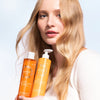 Eine Frau mit langen, gewellten blonden Haaren hält zwei orangefarbene Flaschen Lumene Nordic-C Strength & Shine Shampoo von Lumene vor einem hellblauen und weißen Hintergrund. Sie trägt ein weißes Oberteil und blickt in die Kamera.
