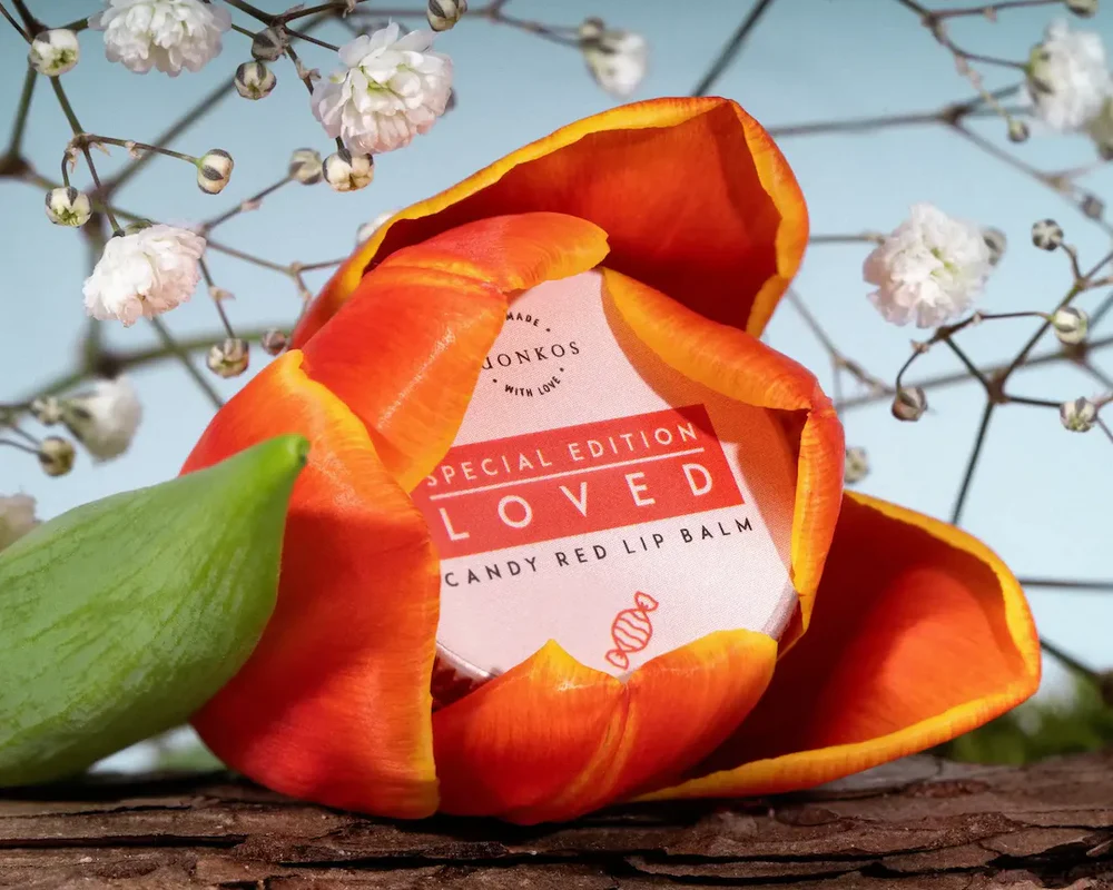 Eine rote und orangefarbene Tulpe umgibt teilweise eine runde Dose mit Luonkos Loved Special Edition Candy Red Lip Balm. Im Hintergrund erscheinen weiße Schleierkrautblüten und eine Holzoberfläche.
