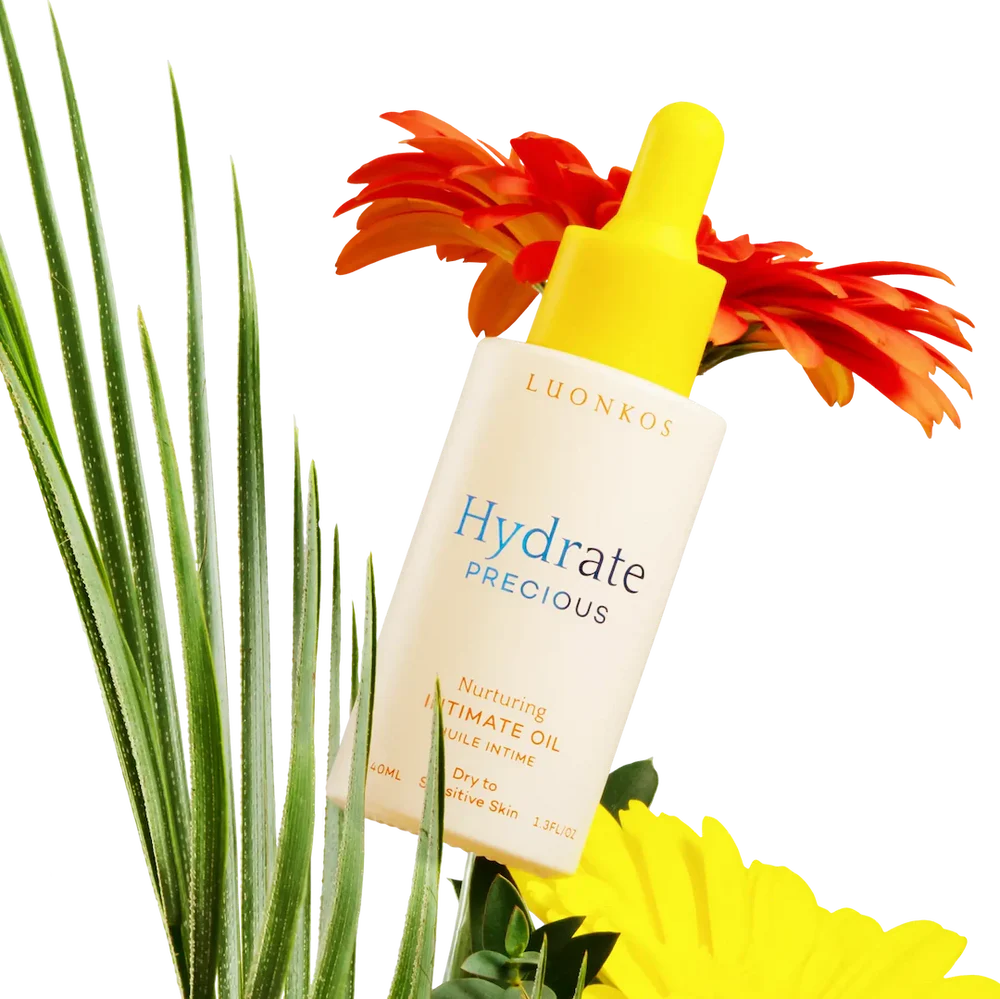 Eine Flasche Luonkos Precious Intimate Oil, ideal für empfindliche und trockene Haut, ruht zwischen grünen Palmenblättern und leuchtend orangefarbenen und gelben Blüten auf weißem Hintergrund.