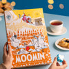 Eine Person hält den Nordqvist Mumin-Adventskalender, eine bunte vegane Teeauswahl von Nordqvist, auf einem blauen Tisch mit einer Tasse Tee, Gebäck und einem blauen herzförmigen Origami. Warme gelbe Lichter leuchten im Hintergrund.