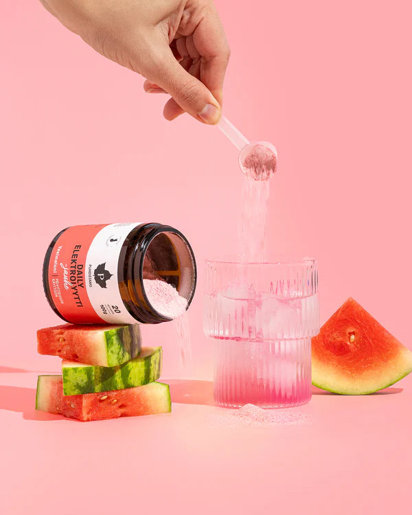 Eine Hand gießt Puhdistamo Daily Electrolyte Powder Watermelon in Wasser neben einem offenen Glas mit der Aufschrift Puhdistamo, das auf Wassermelonenscheiben steht und die tägliche Flüssigkeitszufuhr vor einem rosa Hintergrund hervorhebt.