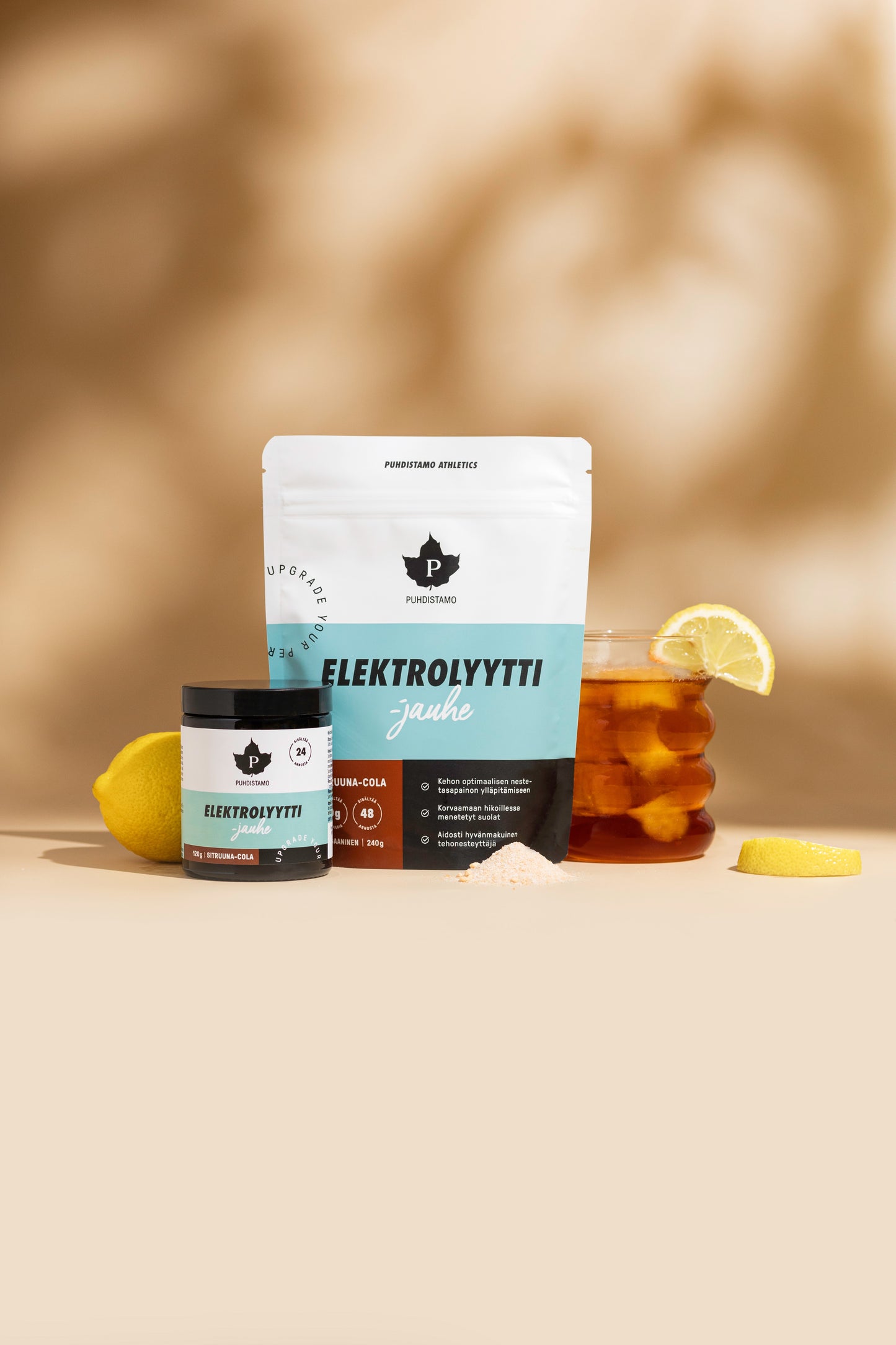 Puhdistamo Electrolyte Powder Lemon & Cola ist mit einem Glas, einem Messlöffel Pulver, einem Glas Eistee mit Zitronenscheibe und Zitronenscheiben auf einer hellbraunen Oberfläche vor einem sanften braunen Hintergrund abgebildet - perfekt für einen hydratisierten und ketogenen Lebensstil.