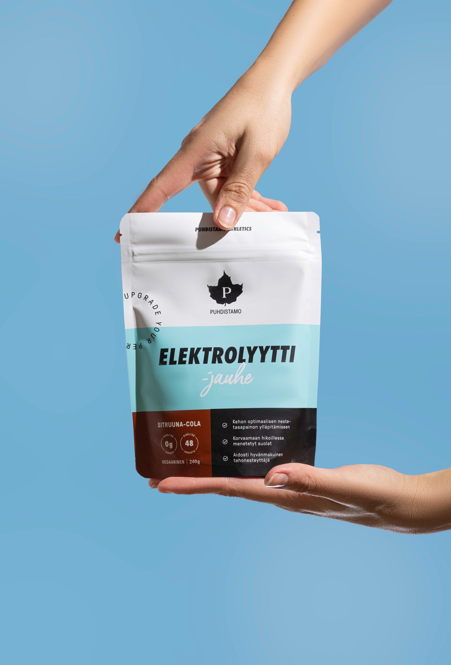 Die Hände halten einen Puhdistamo-Elektrolytpulverbeutel Zitrone & Cola mit der Aufschrift "ELEKTROLYYTTI jauhe" vor einem hellblauen Hintergrund. Die weiß-blaue, ketofreundliche Verpackung zeigt das Puhdistamo-Logo und Angaben zur Flüssigkeitszufuhr.