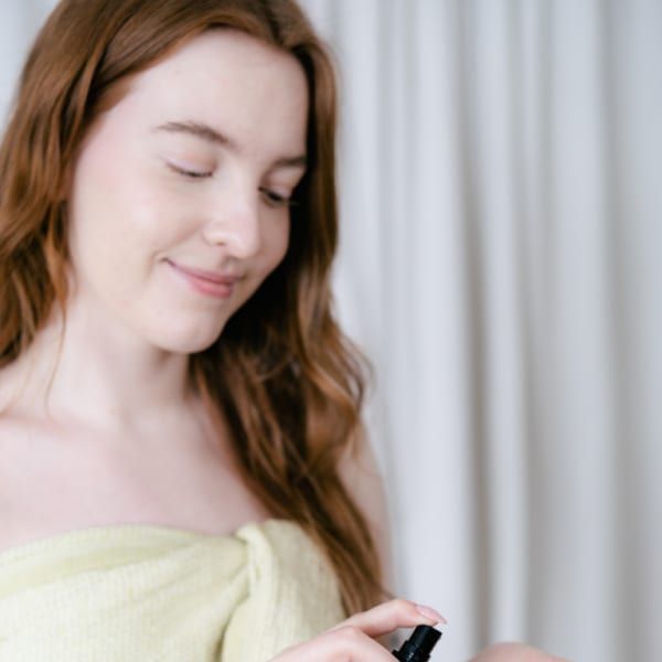 Eine Frau mit langen braunen Haaren, eingewickelt in ein hellgelbes Handtuch, lächelt sanft, während sie das RimitaGold 100% Virgin Argan Oil von Rimita Green aus einer Pumpflasche auf ihre Hand verteilt. Im Hintergrund hängen weiße Vorhänge.