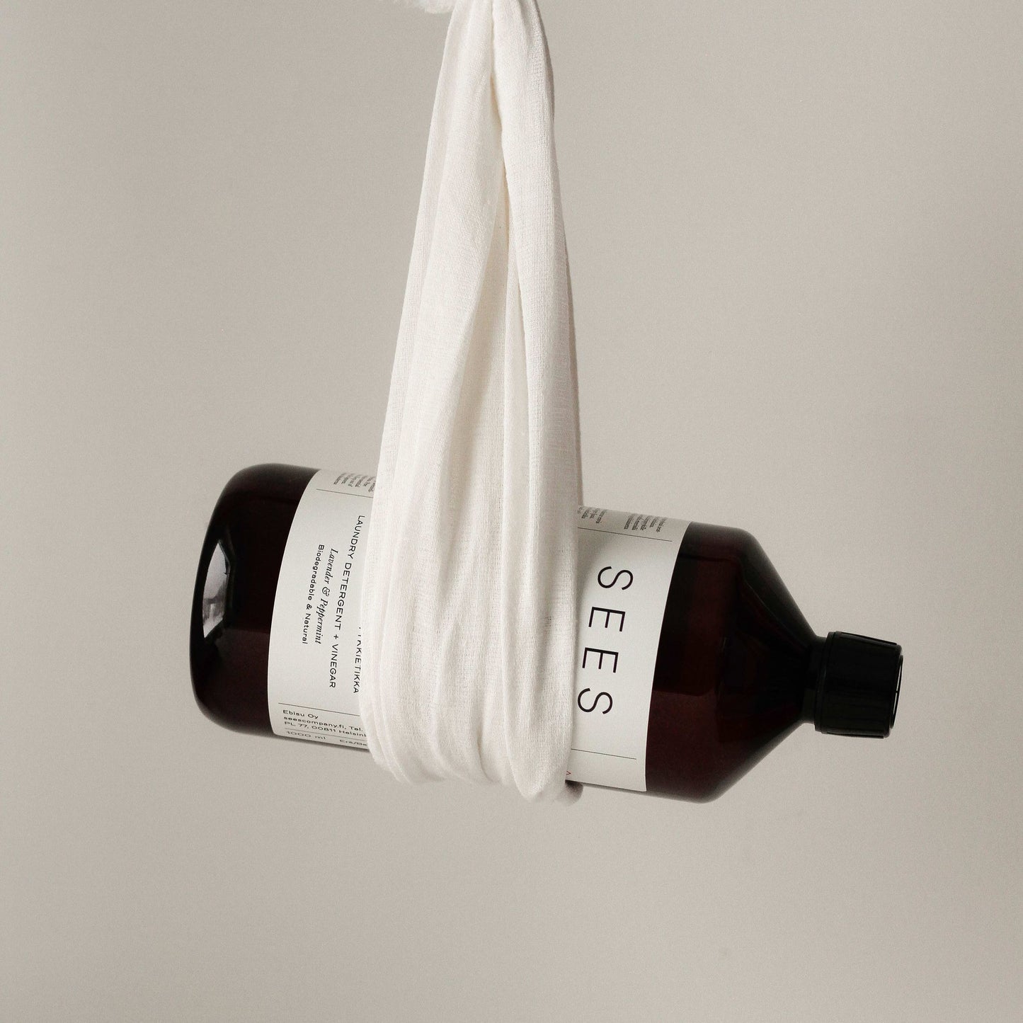 Eine dunkelbraune Flasche SEES Company mit der Aufschrift "SEES 2-in-1 Laundry Detergent with Vinegar" ist in ein weißes Tuch eingewickelt und hängt vor einem schlichten, hellen Hintergrund.