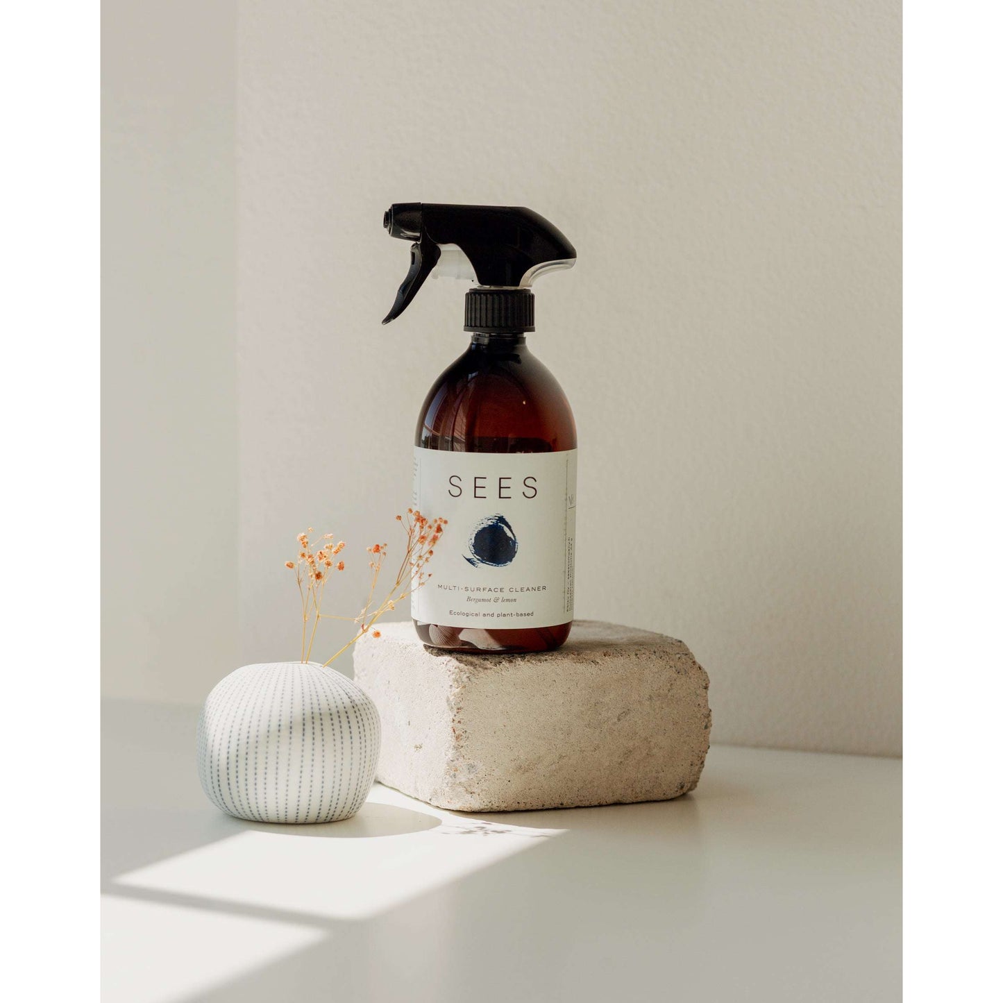 Eine braune Sprühflasche mit dem pflanzlichen Reinigungsspray für Küche und Bad von SEES Companysteht auf einem Steinblock neben einer kleinen weißen Vase mit getrockneten Blumen, die sanft vor einer hellen Wand leuchtet.