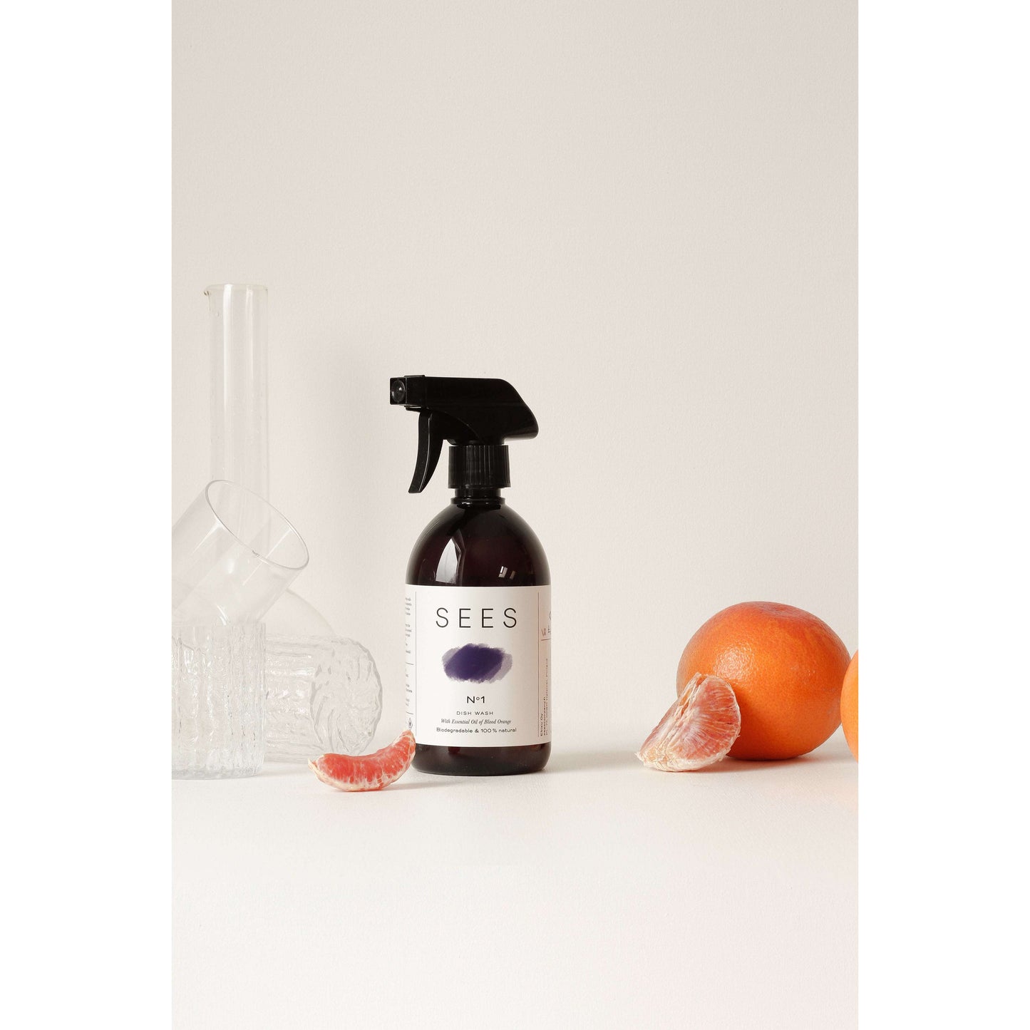 Eine Sprühflasche mit SEES Company steht auf einer weißen Fläche mit Gläsern und Grapefruit und unterstreicht die umweltfreundliche, biologisch abbaubare Reinigung inmitten eines einfachen Lichthintergrunds.