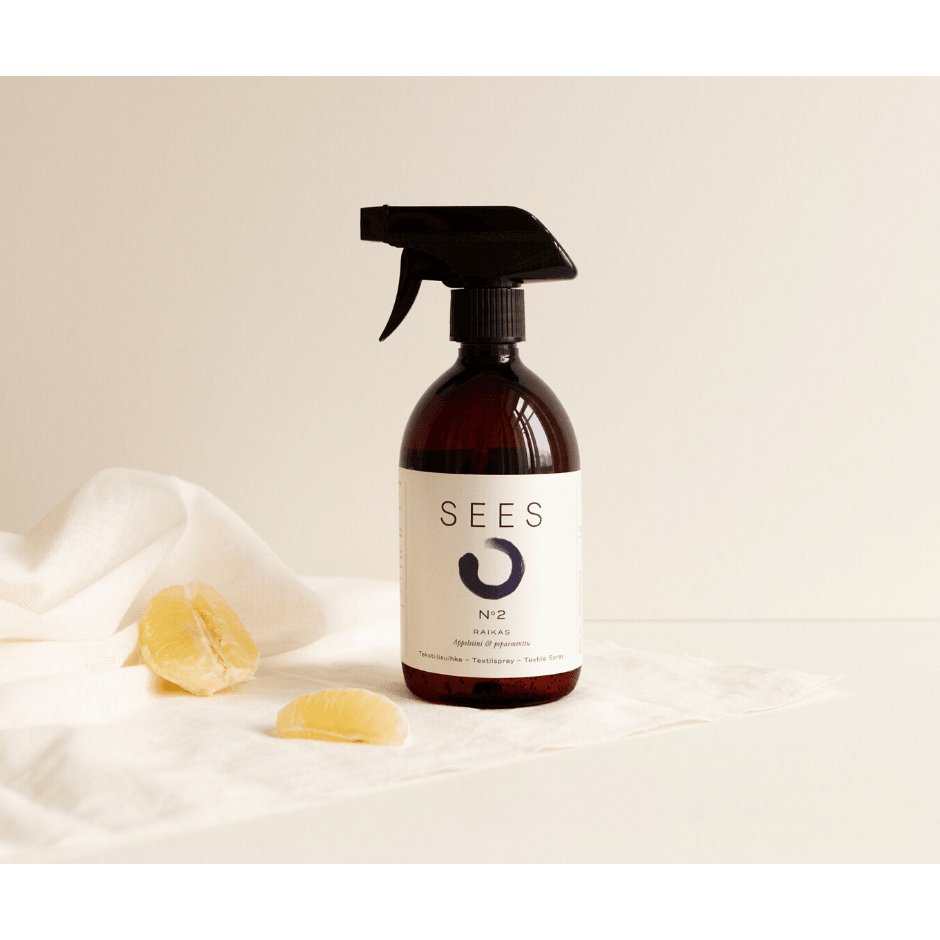Eine dunkelbraune Sprühflasche mit der Aufschrift SEES Textile Spray No. 2 Fresh, Orange & Peppermint von SEES Company steht auf einer weißen Fläche mit einem weißen Tuch und zwei Zitronenscheiben vor einem neutralen, minimalistischen Hintergrund.