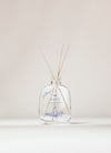 Der Raumduft „Nordic Winter“ von SEES x Moomin Room Diffuser Nordic Winter SEES Company einer klaren Glasflasche mit blauen Pflanzenstoffen und Holzstäbchen, die das finnische Design widerspiegeln, und präsentiert sich elegant vor einem neutralen Hintergrund.