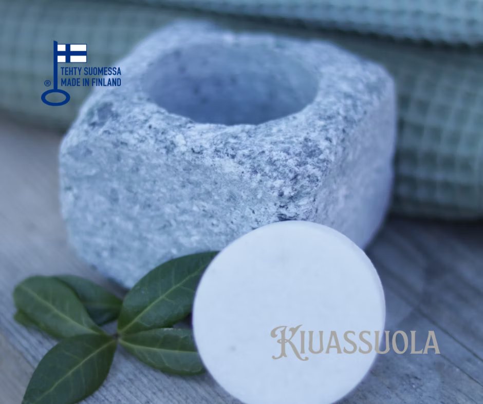 Das Kiuassuola Sauna Salt ILMARI Starter Pack wird in einer Steinschale mit einer weißen Scheibe auf Holz neben grünen Blättern präsentiert. Im Hintergrund erscheint ein grünes Handtuch mit finnischem und englischem Text sowie einer Finnland-Flagge und "KIUASSUOLA".